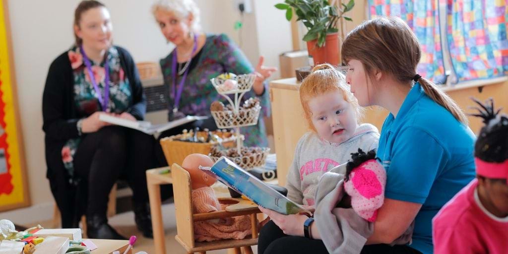 Early Years Practitioner Reading to Child Being Observed by Colleagues in Background